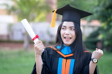Wall Mural - Portrait of a happy and excited young Asian female university graduate wears graduation gown and hat celebrates with a degree in the university campus on the commencement day. Education concept.
