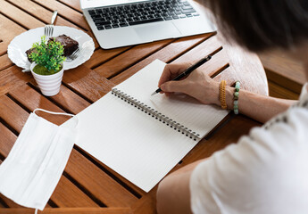 senior asian woman who was retired from work, was writing diary book on the wooden table during covid-19 or coronavirus outbreak. social distancing and new normal concept