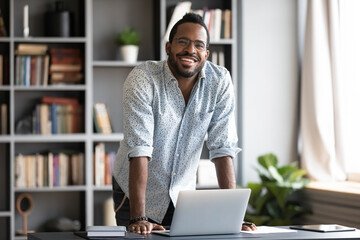 african man in glasses casual shirt leaned over laptop stands in modern office room pose for camera.