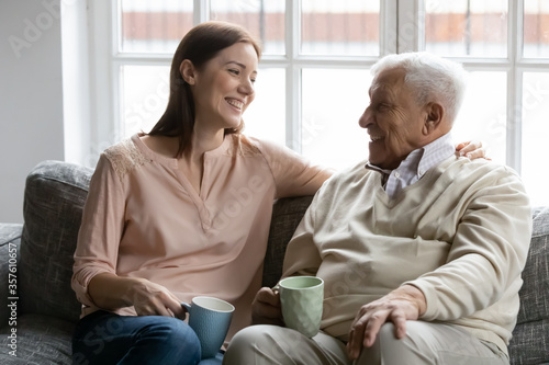 Happy mature man with grownup daughter having fun at home, chatting, talking, sharing news, sitting on cozy couch, smiling older father and young woman drinking tea coffee, enjoying leisure time