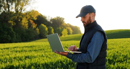 caucasian young male farmer in hat standing in field and typing on keyboard of laptop computer. attr