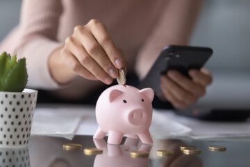 Close up woman holding phone, putting coin in piggy bank, using online banking service apps, saving money, girl planning budget, investment strategy, checking balance, calculating bills