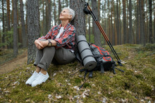 Beautiful Mature Woman In Sneakers And Activewear Sitting On Grass Under Pine Having Rest During Nordic Walking With Sticks And Backpack, Looking Up With Relaxed Carefree Smile, Breathing Fresh Air