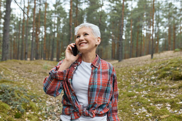 Wall Mural - Cheerful female pensioner with short blonde hair posing in wild nature with pine trees in background, enjoying freshness, sharing impressions with friend, speaking on mobile phone, laughing