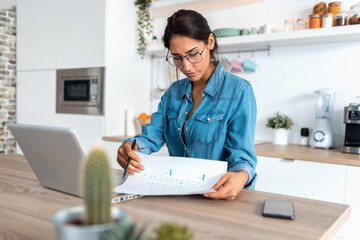 Pretty young woman reviewing paperwork and working on a laptop in the kitchen at home.