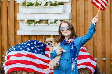 The girl stands in the yard, dressed in a denim dress and holding the flag of USA and little cute dog