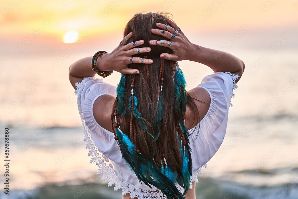 Hippie woman wearing blue feathers in long hair, silver rings with stone and white blouse stands back at sunset. Indie boho vibes and bohemian style - obrazy, fototapety, plakaty 