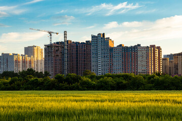 A construction crane on the background of a multi-storey building under construction, in front of which a green field against the setting sky.