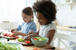 African and Caucasian little girls best friends cooking together in modern kitchen. Multiracial cousins hold knives cutting vegetables on wooden board prepare healthy salad making surprise for parents