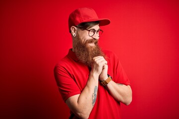 Poster - Young handsome delivery man wearing glasses and red cap over isolated background laughing nervous and excited with hands on chin looking to the side