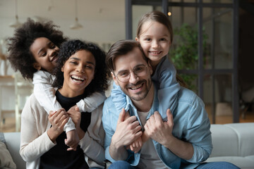 Wall Mural - Cheery African and Caucasian girls cuddle piggy back multi-racial parents loving mom and dad seated on sofa in living room. Happy multinational family portrait, new home funny weekend together concept