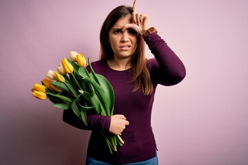 Poster - Young blonde woman holding romantic bouquet of yellow tulips flowers over pink background making fun of people with fingers on forehead doing loser gesture mocking and insulting.