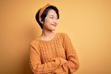 Wall Mural - Young beautiful asian girl wearing casual sweater and diadem standing over yellow background looking to the side with arms crossed convinced and confident