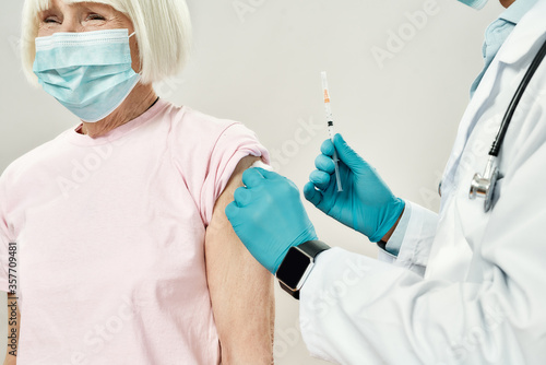 Having an injection. Male doctor in blue sterile gloves holding syringe and making injection to senior woman in medical mask. Vaccination