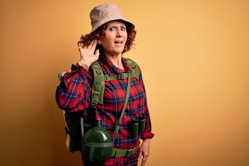 Poster - Middle age curly hair hiker woman hiking wearing backpack and water canteen using binoculars smiling with hand over ear listening an hearing to rumor or gossip. Deafness concept.