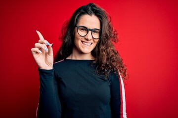 Sticker - Young beautiful woman with curly hair wearing sweater and glasses over red background showing and pointing up with finger number one while smiling confident and happy.