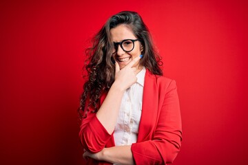 Sticker - Young beautiful woman with curly hair wearing jacket and glasses over red background looking confident at the camera smiling with crossed arms and hand raised on chin. Thinking positive.