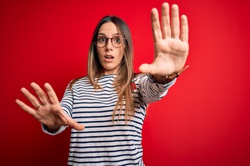 Young beautiful blonde woman with blue eyes wearing glasses standing over red background doing stop gesture with hands palms, angry and frustration expression