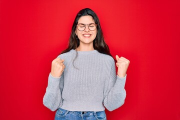 Poster - Young beautiful brunette woman wearing casual sweater over red background excited for success with arms raised and eyes closed celebrating victory smiling. Winner concept.