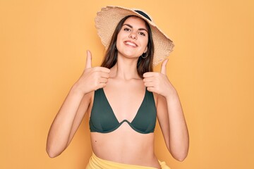 Canvas Print - Young beautiful girl wearing swimwear bikini and summer sun hat over yellow background approving doing positive gesture with hand, thumbs up smiling and happy for success. Winner gesture.