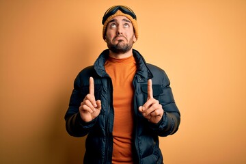 Canvas Print - Young handsome skier man with beard wearing snow sportswear and ski goggles Pointing up looking sad and upset, indicating direction with fingers, unhappy and depressed.