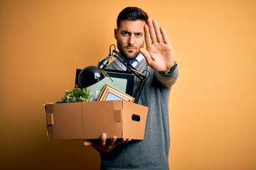 Wall Mural - Young business man holding office box being fired from job over yellow background with open hand doing stop sign with serious and confident expression, defense gesture