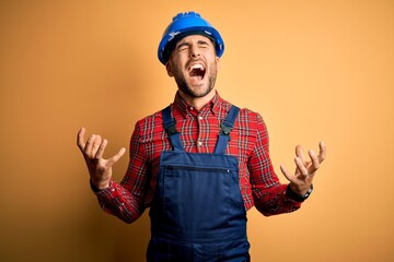 Canvas Print - Young builder man wearing construction uniform and safety helmet over yellow background crazy and mad shouting and yelling with aggressive expression and arms raised. Frustration concept.