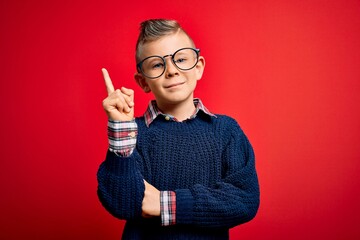 Wall Mural - Young little caucasian kid with blue eyes standing wearing smart glasses over red background with a big smile on face, pointing with hand and finger to the side looking at the camera.