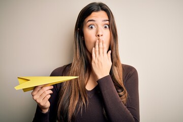 Poster - Young beautiful girl holding paper plane standing over isolated white background cover mouth with hand shocked with shame for mistake, expression of fear, scared in silence, secret concept