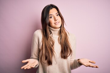 Poster - Young beautiful girl wearing casual turtleneck sweater standing over isolated pink background clueless and confused expression with arms and hands raised. Doubt concept.