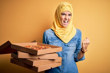 Poster - Young girl wearing muslim hijab holding boxes with Italian pizza over yellow background annoyed and frustrated shouting with anger, crazy and yelling with raised hand, anger concept