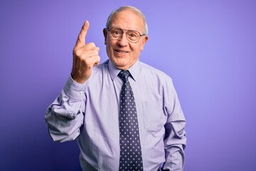 Canvas Print - Grey haired senior business man wearing glasses standing over purple isolated background showing and pointing up with finger number one while smiling confident and happy.