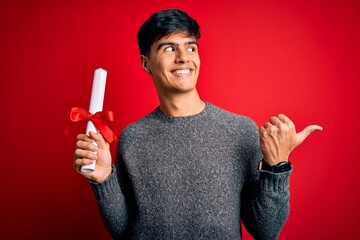 Poster - Young student man holding university graduated diploma degree over red background pointing and showing with thumb up to the side with happy face smiling