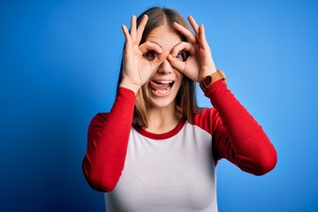 Wall Mural - Young beautiful redhead woman wearing casual t-shirt over isolated blue background doing ok gesture like binoculars sticking tongue out, eyes looking through fingers. Crazy expression.