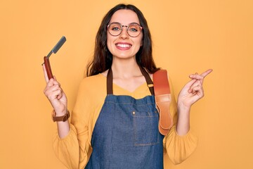 Wall Mural - Young beautiful barber woman wearing apron holding razor blade over yellow background very happy pointing with hand and finger to the side