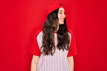 Wall Mural - Beautiful woman with blue eyes wearing baseball sportswear and cap over red background looking away to side with smile on face, natural expression. Laughing confident.