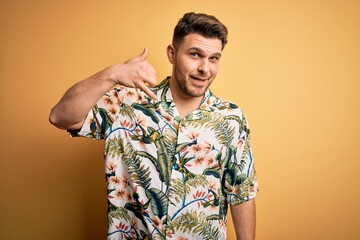 Wall Mural - Young man with blue eyes on vacation wearing floral summer shirt over yellow background smiling doing phone gesture with hand and fingers like talking on the telephone. Communicating concepts.