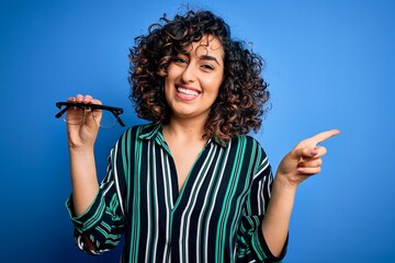 Poster - Young beautiful optical arab woman holding vision glasses over isolated blue background very happy pointing with hand and finger to the side