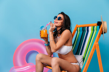 Wall Mural - Appealing tanned girl sitting on deck chair and drinking cocktail. Studio shot of lady in sunglasses posing on chaise longue on blue background.