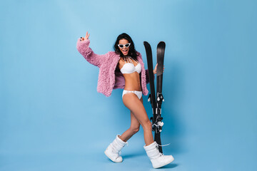 Wall Mural - Blithesome brunette girl in bikini and jacket dancing on blue background. Full length view of pretty young woman with skis.