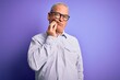 © Krakenimages.com - Middle age handsome hoary man wearing striped shirt and glasses over purple background touching mouth with hand with painful expression because of toothache or dental illness on teeth. Dentist