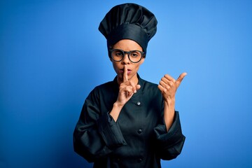 Wall Mural - Young african american chef woman wearing cooker uniform and hat over blue background asking to be quiet with finger on lips pointing with hand to the side. Silence and secret concept.