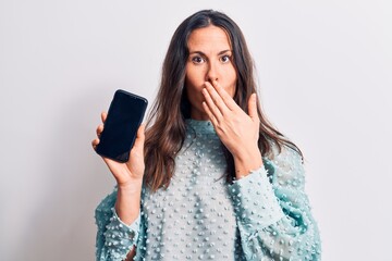 Young beautiful brunette woman holding smartphone showing screen over white background covering mouth with hand, shocked and afraid for mistake. Surprised expression