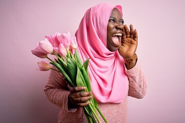 Poster - Young african american plus size woman wearing muslim hijab holding bouquet of pink tulips shouting and screaming loud to side with hand on mouth. Communication concept.