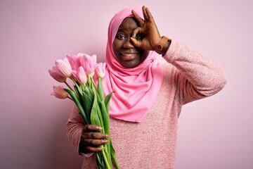 Poster - Young african american plus size woman wearing muslim hijab holding bouquet of pink tulips doing ok gesture with hand smiling, eye looking through fingers with happy face.