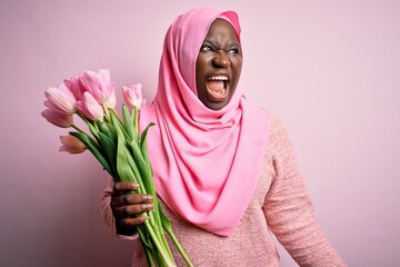 Poster - Young african american plus size woman wearing muslim hijab holding bouquet of pink tulips angry and mad screaming frustrated and furious, shouting with anger. Rage and aggressive concept.