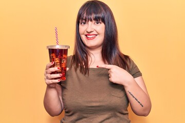 Young plus size woman drinking glass of cola beverage smiling happy pointing with hand and finger