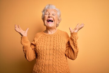 Wall Mural - Senior beautiful grey-haired woman wearing casual sweater over isolated yellow background celebrating mad and crazy for success with arms raised and closed eyes screaming excited. Winner concept