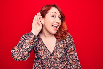 Sticker - Young beautiful redhead woman wearing floral casual dress standing over red background smiling with hand over ear listening and hearing to rumor or gossip. Deafness concept.