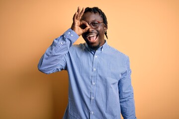 Sticker - Young handsome african american man wearing shirt and glasses over yellow background doing ok gesture with hand smiling, eye looking through fingers with happy face.